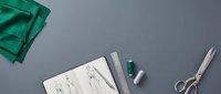 Fashion designer sketches with green fabric swatch, measuring tape, scissors and pencils on gray background for clothing design, tailoring and fashion illustration projects.