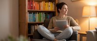 Young woman relaxing at home with laptop computer in cozy living room with colorful bookshelf and warm lighting for remote work or online learning.