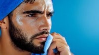 Athletic man with beard wearing blue beanie against vibrant blue background, wiping sweat with towel after workout, showing determination and focus in fitness portrait.