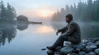 Peaceful morning fishing scene with angler sitting on rocky shore of misty lake at sunrise surrounded by pine forest and cabin in background.
