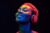 Young African woman with short pink hair wearing headphones, smiling with eyes closed in neon lighting against dark background, expressing joy and musical enjoyment.