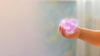 Woman's hand holding glowing neon heart symbol against blurred urban background, representing love, care, charity, and emotional connection for healthcare and wellness concepts.
