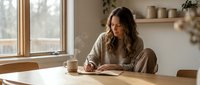 Young woman writing in journal at wooden table by window in cozy home interior with natural light and plants creating peaceful morning atmosphere.