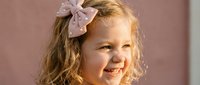 Young blonde girl with curly hair wearing pink bow smiling joyfully in warm sunlight. Cheerful child portrait for family lifestyle and childhood concepts.