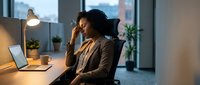 Stressed African American businesswoman working late at office desk, experiencing headache while using laptop computer in modern workplace during evening hours.