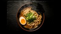 Steaming hot ramen noodles in dark bowl with soft-boiled egg, green onions and rich broth on slate background for restaurant menu design.