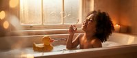 Young African American woman enjoying relaxing bubble bath in modern bathroom with warm golden lighting and candles for spa wellness routine.