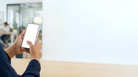 Person with dark skin tone holding smartphone with blank white screen at wooden table, office environment background.