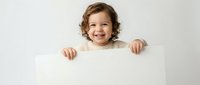 Happy toddler boy with curly brown hair holding blank white sign for text, smiling child in beige sweater on neutral background for advertising mockup.