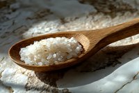 Coarse sea salt crystals on wooden spoon against white textured background, natural lighting creates soft shadows highlighting mineral texture and purity.
