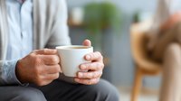 Person holding white coffee mug with both hands while sitting in cozy living room, creating warm atmosphere of relaxation and comfort.