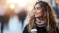 Young woman with long curly hair wearing winter sweater and knit scarf holds coffee cup while smiling outdoors during golden sunset with bokeh city lights background.