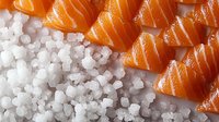 Fresh raw salmon sashimi slices arranged diagonally on coarse sea salt crystals, creating contrast between orange fish and white mineral texture.