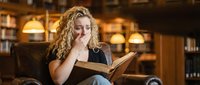 Young blonde woman with curly hair reading book in cozy library setting with warm lighting and wooden shelves in background.
