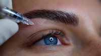Close-up of cosmetic eyebrow microblading procedure with gloved hand holding needle tool near woman's blue eye and defined brow during beauty treatment.