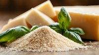 Fresh grated parmesan cheese pile with whole cheese blocks and green basil leaves on wooden surface in natural lighting, macro food photography.