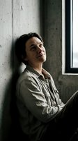 Thoughtful middle-aged man in casual shirt sitting by concrete wall and window, looking up with hopeful expression in modern industrial interior space.