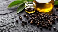 Black peppercorns scattered on dark stone surface with glass jar of olive oil and green leaves, dramatic side lighting creates gourmet food atmosphere.