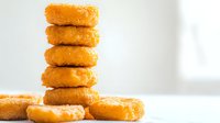 Golden crispy chicken nuggets stacked in tower formation on white surface, close-up view showing breaded texture and warm appetizing color against bright background.