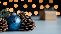 Festive blue glitter Christmas bauble with natural pine cones against blurred warm bokeh lights background, creating magical holiday atmosphere.