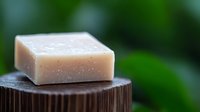 Natural handmade soap bar on wooden surface against blurred green background, organic skincare product with gentle texture and soft beige color.