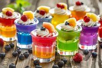 Colorful layered fruit jelly desserts in shot glasses topped with whipped cream and fresh berries on rustic wooden table.