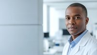 Professional African American male doctor in white lab coat smiling confidently in modern medical facility with clean laboratory background for healthcare concepts.