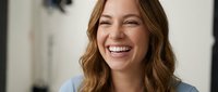 Young Caucasian woman with wavy brown hair laughing joyfully in bright modern interior, expressing genuine happiness and positive emotions for lifestyle content.
