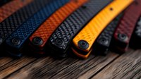 Colorful collection of folding knives with textured handles in black, orange, and burgundy arranged in row on rustic wooden surface with dramatic lighting.