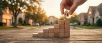 Hand placing wooden block on staircase stack with suburban houses blurred in background, representing real estate growth, property investment, and housing market development.