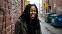 Young African American woman with braided hair smiling joyfully against brick wall in urban alley. Authentic portrait for lifestyle and diversity campaigns.