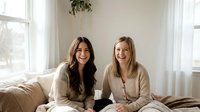 Two happy young women friends sitting together on cozy bed in bright bedroom laughing and bonding during casual home visit or sleepover gathering.