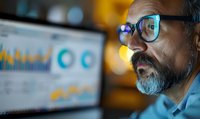 Middle-aged businessman with gray beard analyzing financial data on computer screen with colorful charts and graphs in evening office environment.