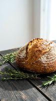 Freshly baked artisan sourdough bread loaf with golden crust on rustic wooden table with rosemary herbs for homemade bakery and cooking concepts.