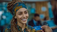 Young African woman with dreadlocks wearing traditional colorful headwrap and clothing, smiling while holding blue credit card in modern office setting.