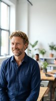 Mature Caucasian businessman with curly hair smiling confidently in modern office workspace with natural light and plants for professional success concepts.