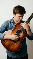 Young male musician with dark hair and Caucasian ethnicity playing acoustic guitar while wearing denim shirt. Suitable for music education, lessons, talent, and performance concepts.