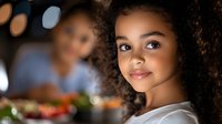 Young African American girl with curly natural hair smiling warmly at dinner table with fresh healthy meal and family gathering in background, natural lighting creates cozy atmosphere.