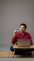 Young Indian businessman in red shirt pointing up with eureka moment while working on laptop at wooden desk for business presentations and startup concepts.