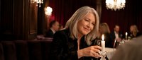 Elegant mature Caucasian woman with long blonde hair enjoying wine at upscale restaurant dinner party with warm ambient lighting and crystal chandeliers.