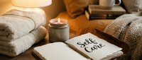 Cozy self care setup with open journal, lit candle, folded towels, and warm blanket on bedside table in soft golden light.
