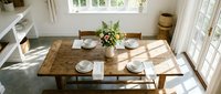 Rustic wooden dining table set for four with white plates and fresh flowers in bright sunlit room with natural light streaming through windows.