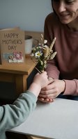 Child giving wildflower bouquet to smiling mother on Mother's Day with greeting card and coffee mug on wooden table in cozy home setting.