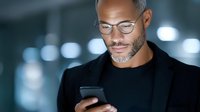 Professional African American businessman with glasses using smartphone in modern office environment with bokeh lighting effects.
