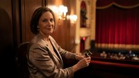 Elegant mature businesswoman in beige suit smiling confidently in luxurious theater box with red velvet curtains and warm lighting for corporate events.