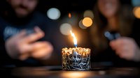Single burning candle with ornate silver holder glowing against dark background with blurred bokeh lights and silhouettes, creating intimate celebratory atmosphere.