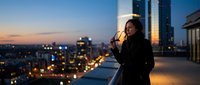 Woman with dark hair and medium skin tone in professional black blazer holding coffee cup on rooftop terrace at dusk, overlooking illuminated cityscape at golden hour for corporate lifestyle and business themes.