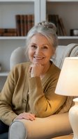 Senior woman with gray hair smiling at home, wearing mustard cardigan, sitting on couch with lamp and bookshelf background, portrait for lifestyle and aging content.