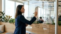 Professional businesswoman writing on glass board with development pipeline and innovation strategy in modern office environment.