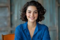 Young Mediterranean woman with curly dark hair and bright smile wearing blue business attire, sitting in modern office setting against blurred background.